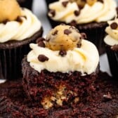 Chocolate cupcake with white frosting, topped with a cookie dough ball and chocolate chips. The cupcake has a bite taken out, revealing cookie dough inside. Other cupcakes are visible in the background.