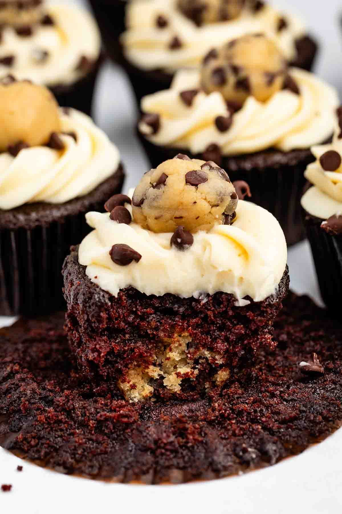 Chocolate cupcake with white frosting, topped with a cookie dough ball and chocolate chips. The cupcake has a bite taken out, revealing cookie dough inside. Other cupcakes are visible in the background.