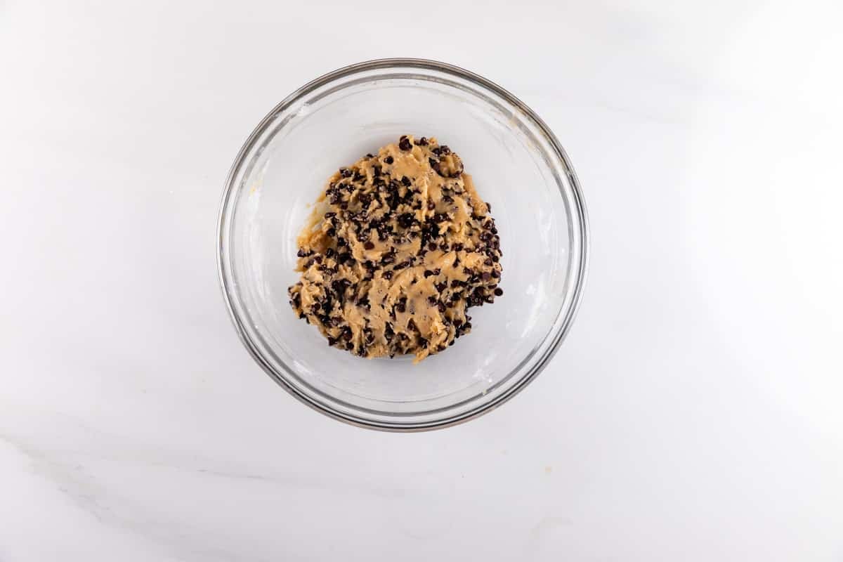 A clear glass bowl filled with chocolate chip cookie dough sits on a white marble surface.