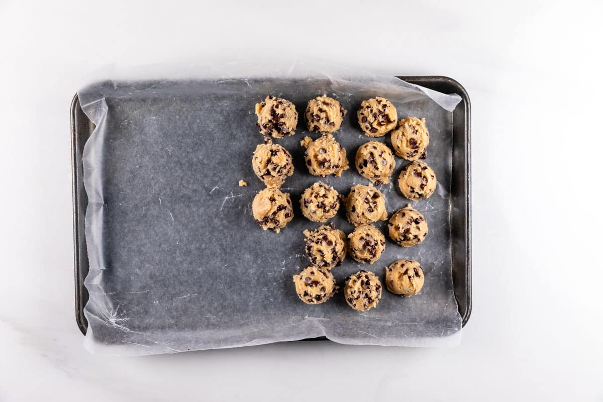 A baking tray lined with parchment paper holds 16 raw chocolate chip cookie dough balls arranged on the right half, ready to be baked.