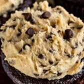A close-up of a chocolate cupcake topped with a generous scoop of cookie dough and chocolate chips, with another cupcake blurred in the background.