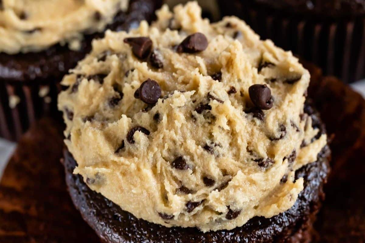 A close-up of a chocolate cupcake topped with a generous scoop of cookie dough and chocolate chips, with another cupcake blurred in the background.