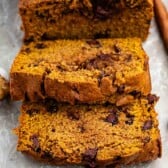 Sliced pumpkin bread with chocolate chips on a tray, surrounded by two cinnamon sticks, a spoonful of peanut butter, and two small pumpkin-shaped candies.