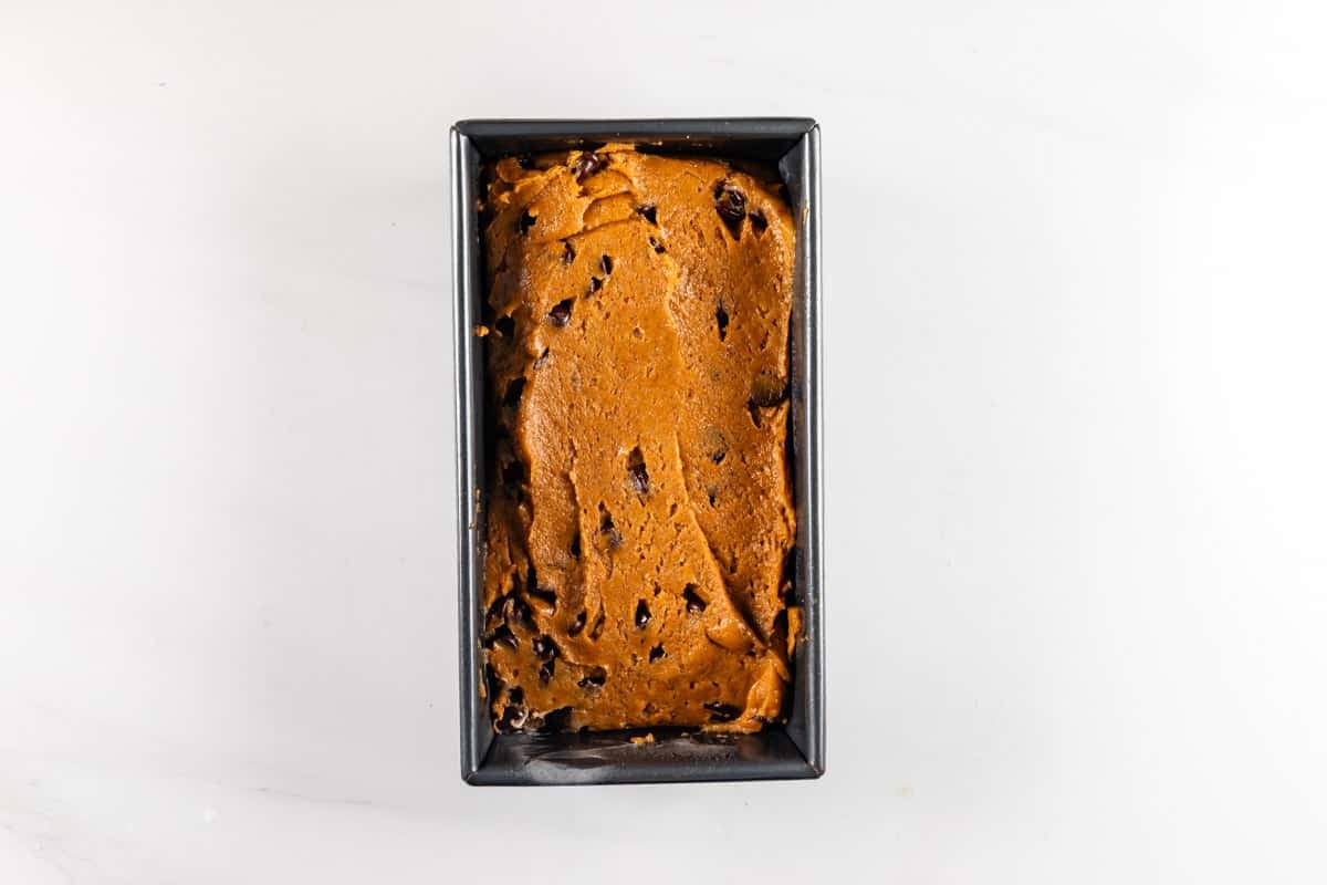 Rectangular loaf pan filled with unbaked brown batter containing dark chocolate chunks, sitting on a white surface.