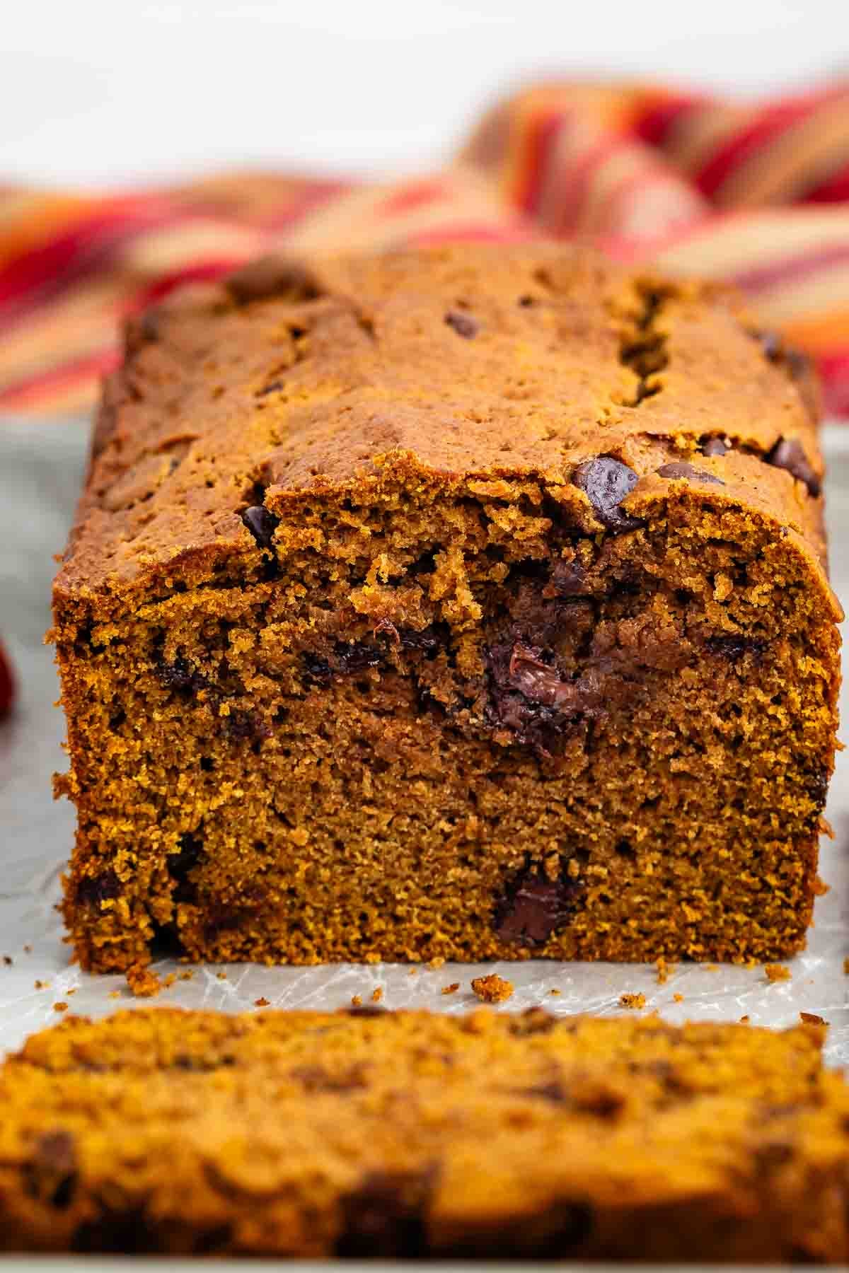 A close-up of a sliced loaf of pumpkin bread with chocolate chips on a white surface, with a colorful striped cloth in the background.