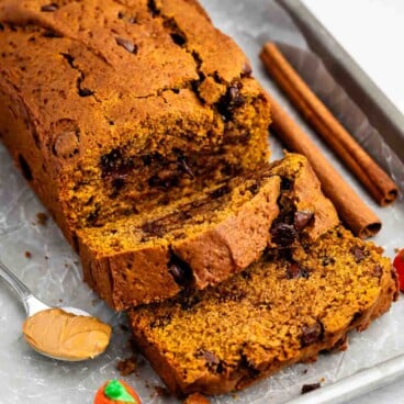 Sliced pumpkin bread with chocolate chips on a tray, a spoonful of peanut butter, two cinnamon sticks, and a small decorative candy pumpkin beside it.