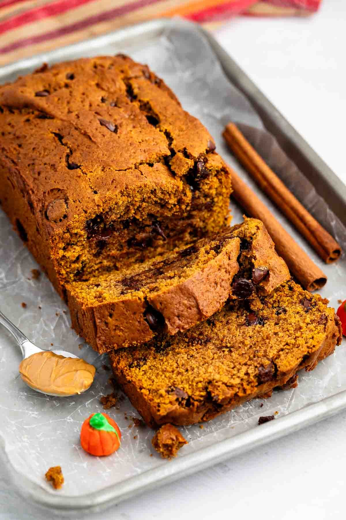 Sliced pumpkin bread with chocolate chips on a tray, a spoonful of peanut butter, two cinnamon sticks, and a small decorative candy pumpkin beside it.