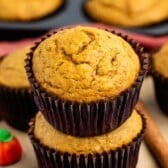 Two pumpkin muffins stacked, with a small candy pumpkin and a cinnamon stick nearby. In the background, more muffins sit in a pan, all on a wooden surface.