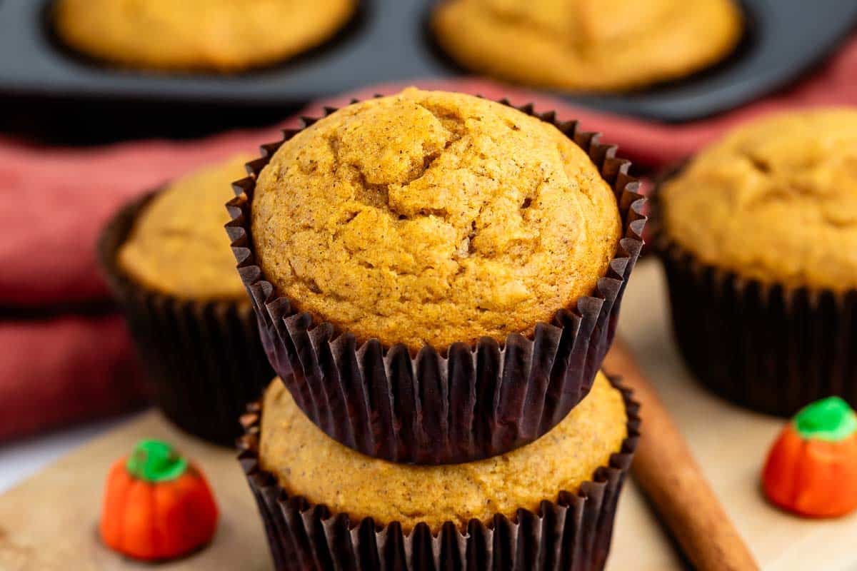 Two pumpkin muffins stacked, with a small candy pumpkin and a cinnamon stick nearby. In the background, more muffins sit in a pan, all on a wooden surface.