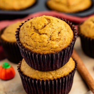 Two pumpkin muffins stacked on a wooden board with a candy pumpkin and cinnamon stick beside them; more muffins are in a baking tray in the background.