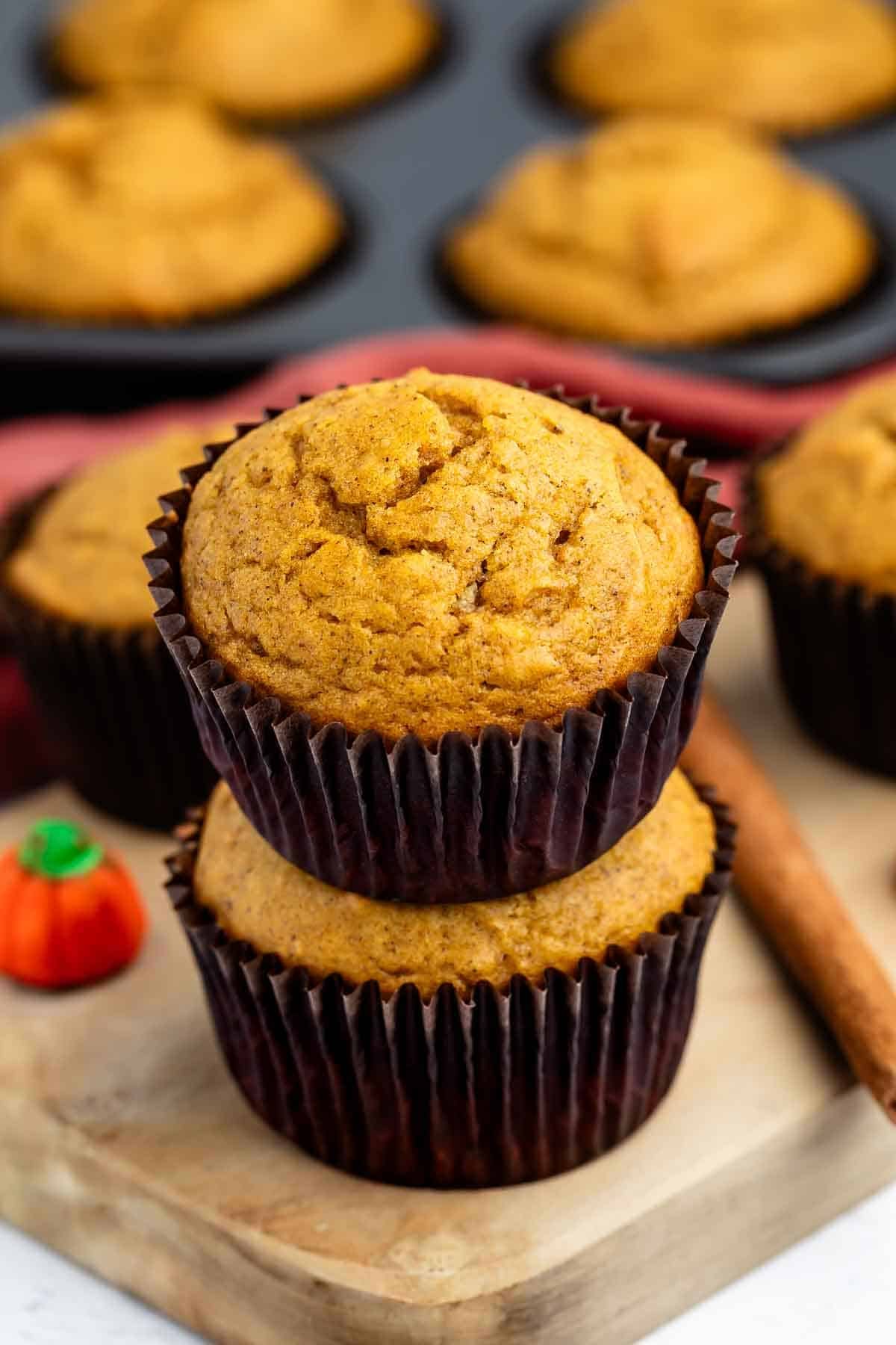 Two pumpkin muffins stacked on a wooden board with a candy pumpkin and cinnamon stick beside them; more muffins are in a baking tray in the background.