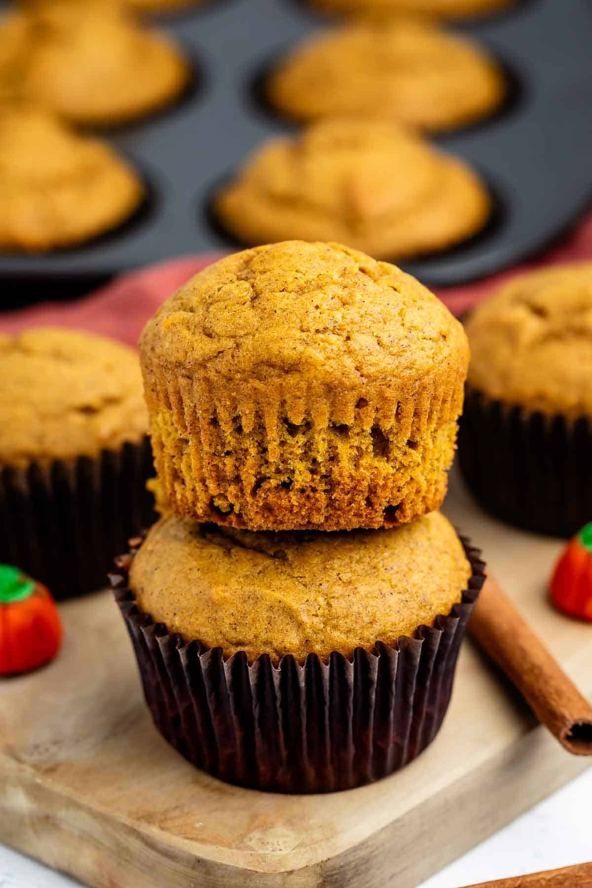 Two pumpkin muffins stacked on a wooden board, with a cinnamon stick and small pumpkin-shaped candies nearby. A muffin tray with more muffins is blurred in the background.