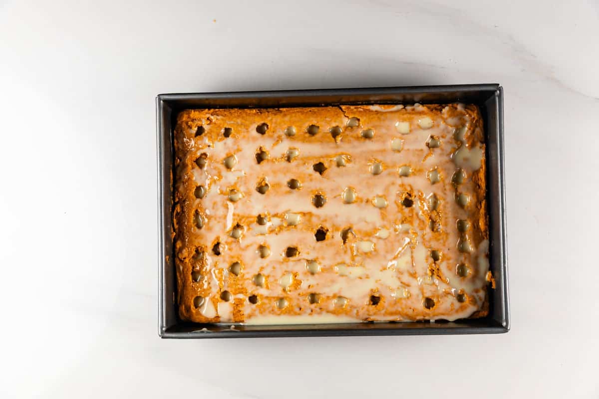 A rectangular cake with evenly spaced holes filled with glaze sits in a black baking pan on a white surface.