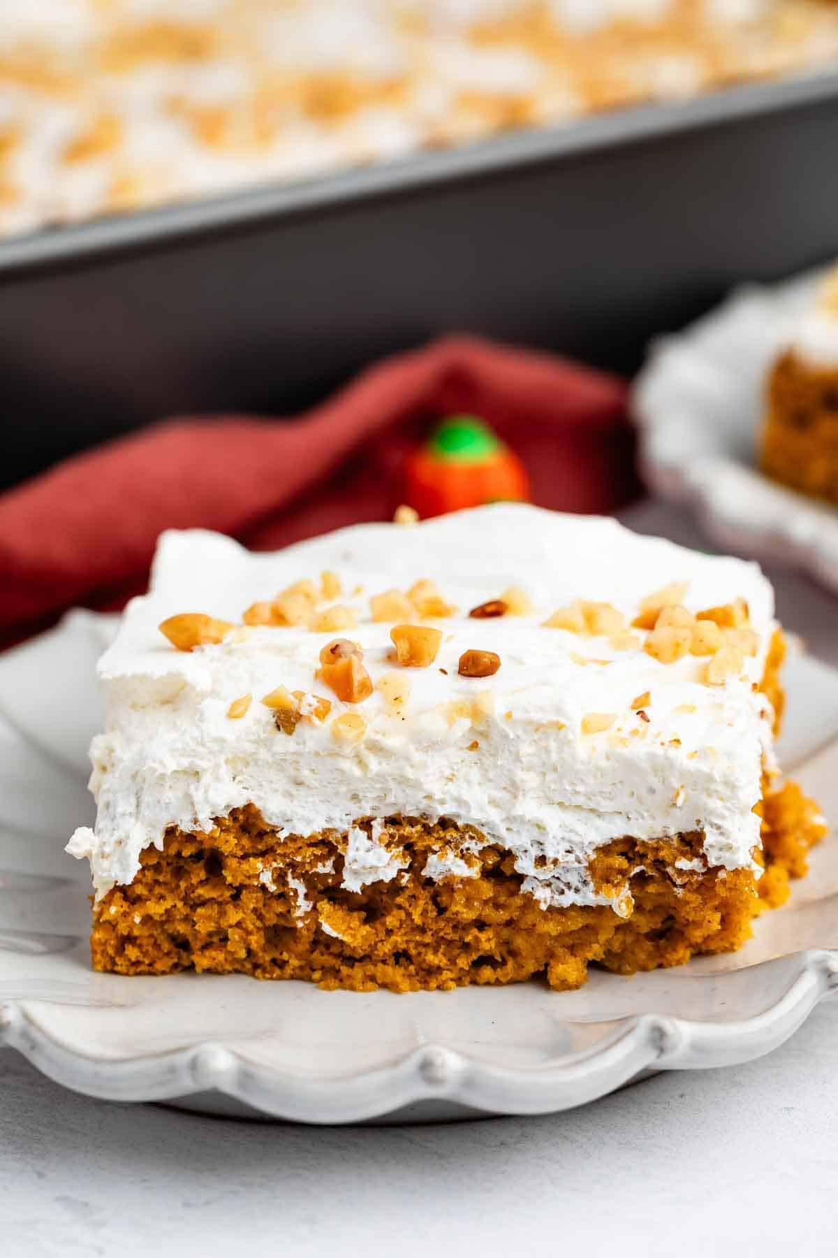 A square piece of pumpkin cake with white frosting and chopped nuts on top sits on a decorative gray plate.