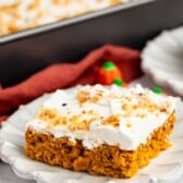 A square piece of pumpkin cake with white frosting and chopped nuts on top sits on a decorative white plate, with a pan of more cake in the background.
