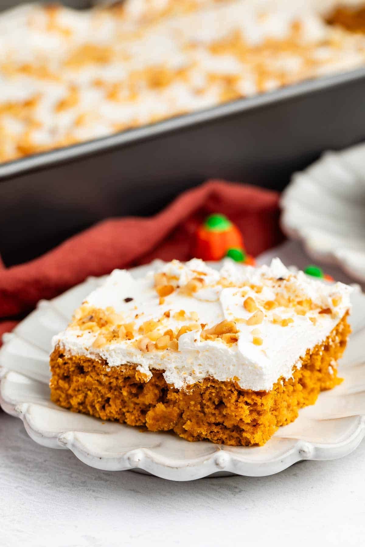 A square piece of pumpkin cake with white frosting and chopped nuts on top sits on a decorative white plate, with a pan of more cake in the background.