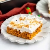 A slice of pumpkin cake with white frosting and chopped nuts on a scalloped plate, next to a gold fork and small pumpkin candies.