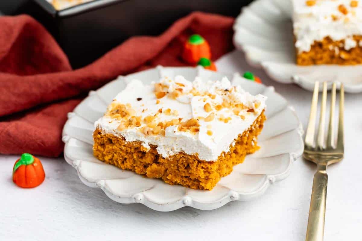 A slice of pumpkin cake with white frosting and chopped nuts on a scalloped plate, next to a gold fork and small pumpkin candies.
