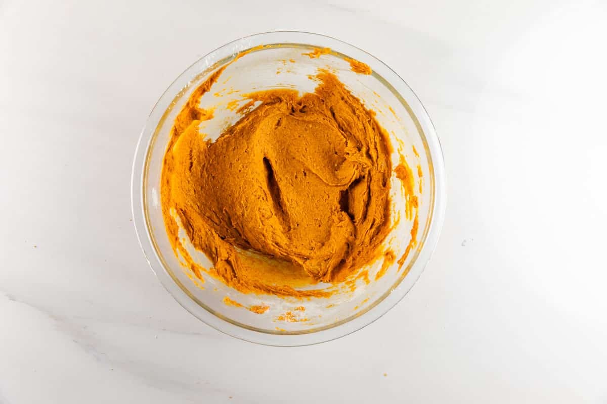 A glass bowl filled with orange cookie dough sits on a white surface. The dough has been partially mixed, leaving swirls and some dough sticking to the sides of the bowl.