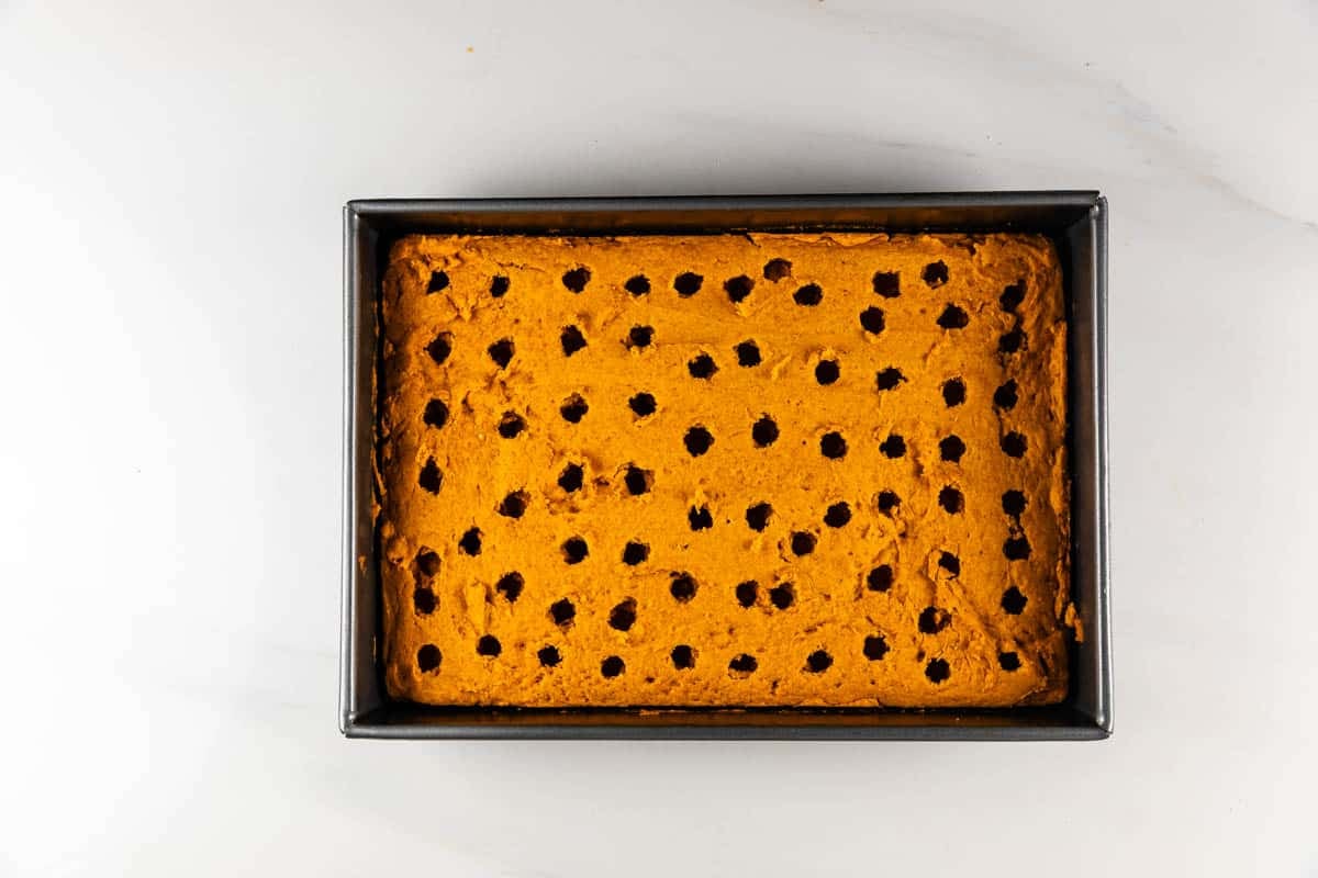 A rectangular pan holds an orange cake with evenly spaced holes poked into the surface, placed on a white countertop.