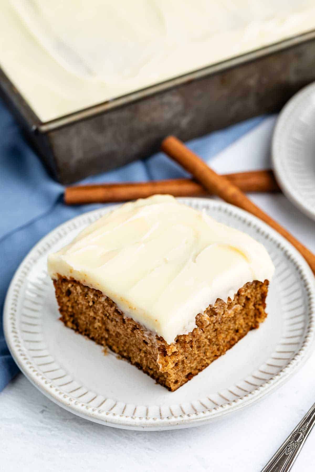 A square slice of spiced cake with white frosting sits on a plate, with cinnamon sticks and the rest of the cake in a pan in the background.