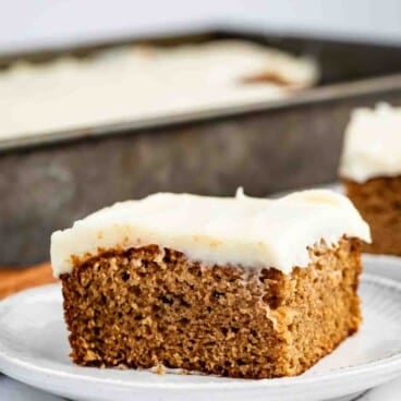 A slice of frosted spice cake sits on a white plate, with the rest of the cake visible in a baking pan in the background.