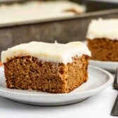 A slice of frosted spice cake sits on a white plate, with another slice and a fork in the background. The cake looks moist and is topped with a thick layer of creamy white frosting.