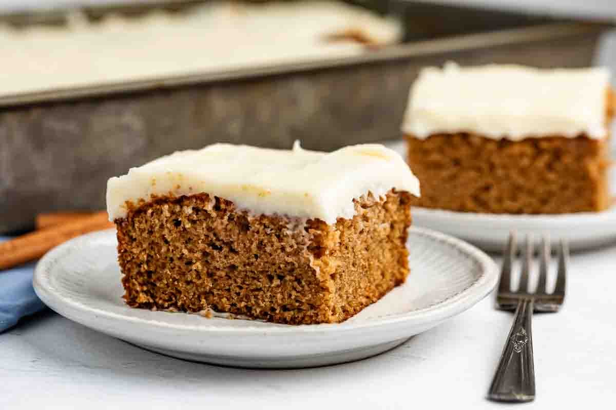 A slice of frosted spice cake sits on a white plate, with another slice and a fork in the background. The cake looks moist and is topped with a thick layer of creamy white frosting.