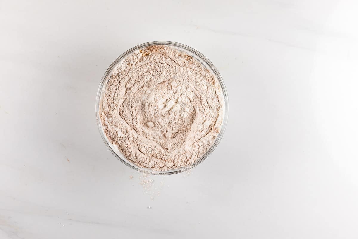 A clear glass bowl filled with flour sits on a white marble surface, with some flour scattered around the bowl.