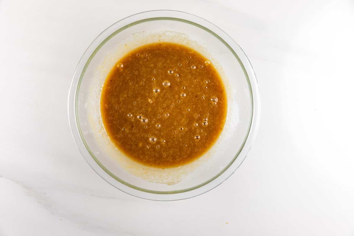 A glass bowl filled with a brown liquid mixture, possibly batter or sauce, sits on a white marble surface.