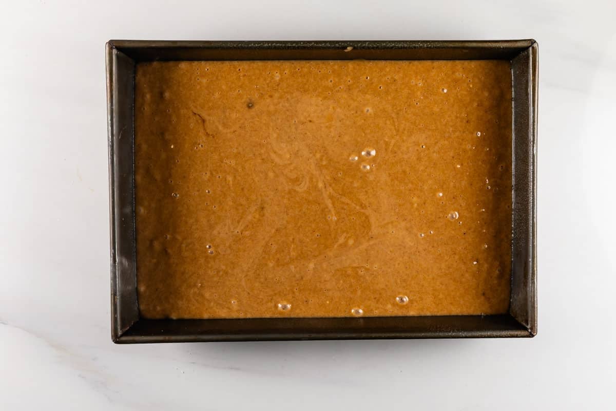 A rectangular metal baking pan filled with light brown cake batter, ready to be baked, is placed on a white countertop.