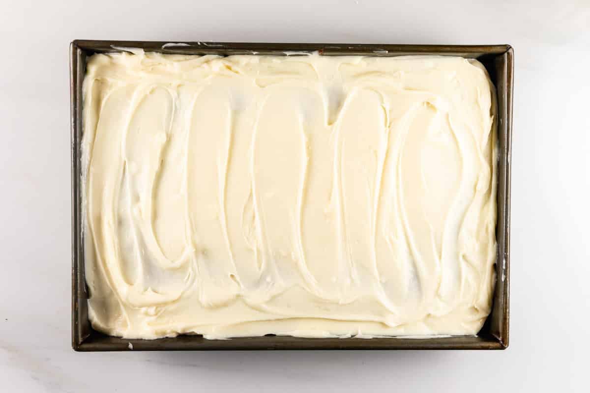 A rectangular cake in a metal pan is covered with a thick layer of creamy white frosting, smoothly spread with visible swirls.