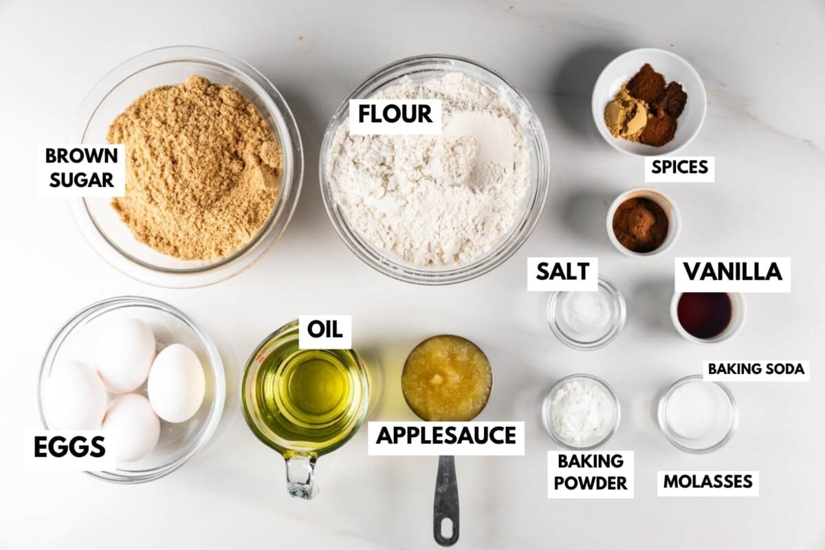 Various baking ingredients in bowls on a white surface, including labeled brown sugar, flour, eggs, oil, applesauce, spices, salt, vanilla, baking soda, baking powder, and molasses.