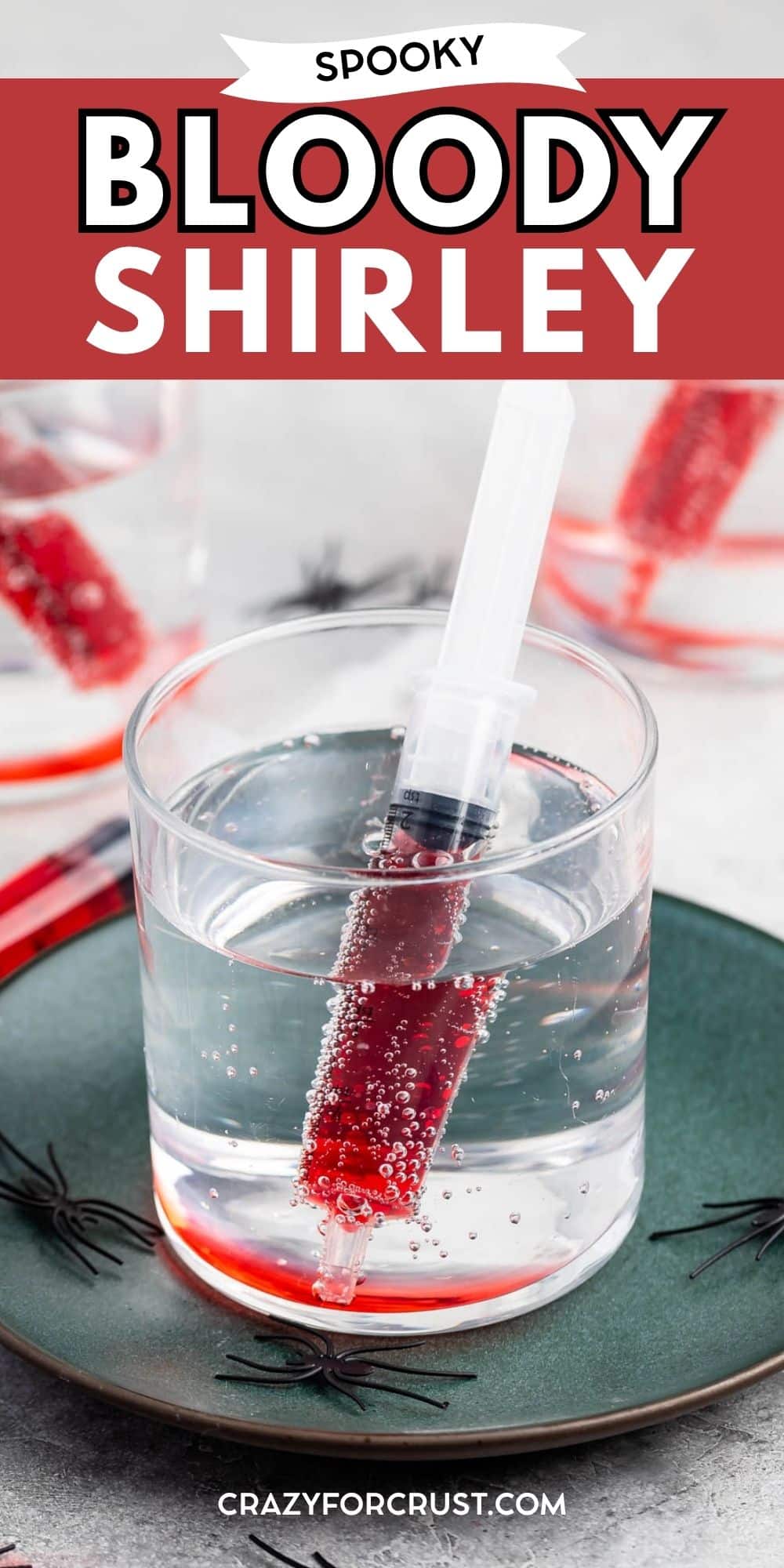 A clear drink in a glass with a red-filled syringe inside, sitting on a green plate. Black plastic spiders decorate the plate. The image text reads Spooky Bloody Shirley..