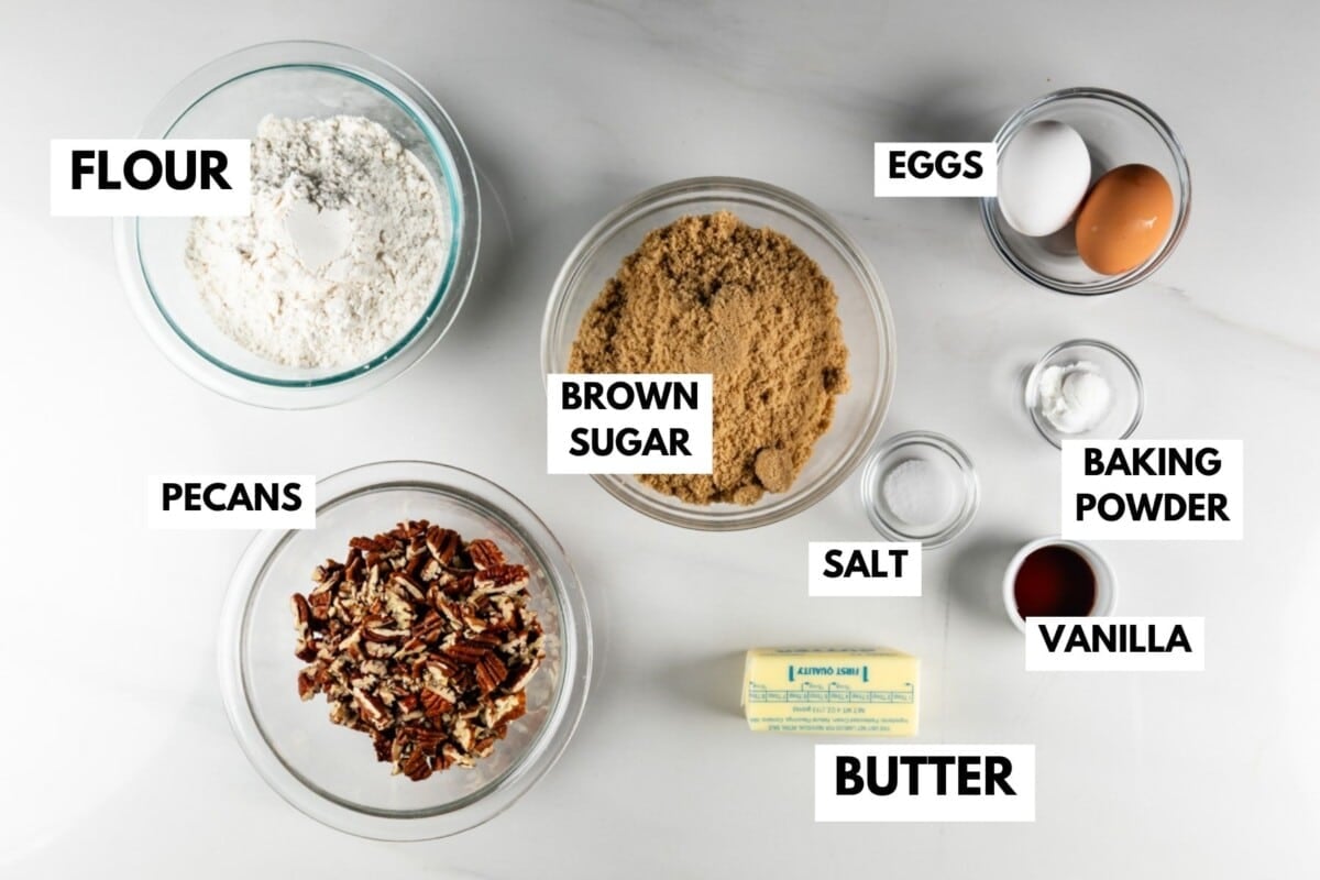 Labeled baking ingredients on a white surface: flour, brown sugar, eggs, pecans, butter, salt, baking powder, and vanilla in bowls or containers.