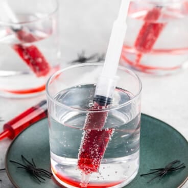 A clear drink with a syringe filled with red liquid inside the glass, served on a green plate with small plastic black spiders, and two more similar drinks in the background.