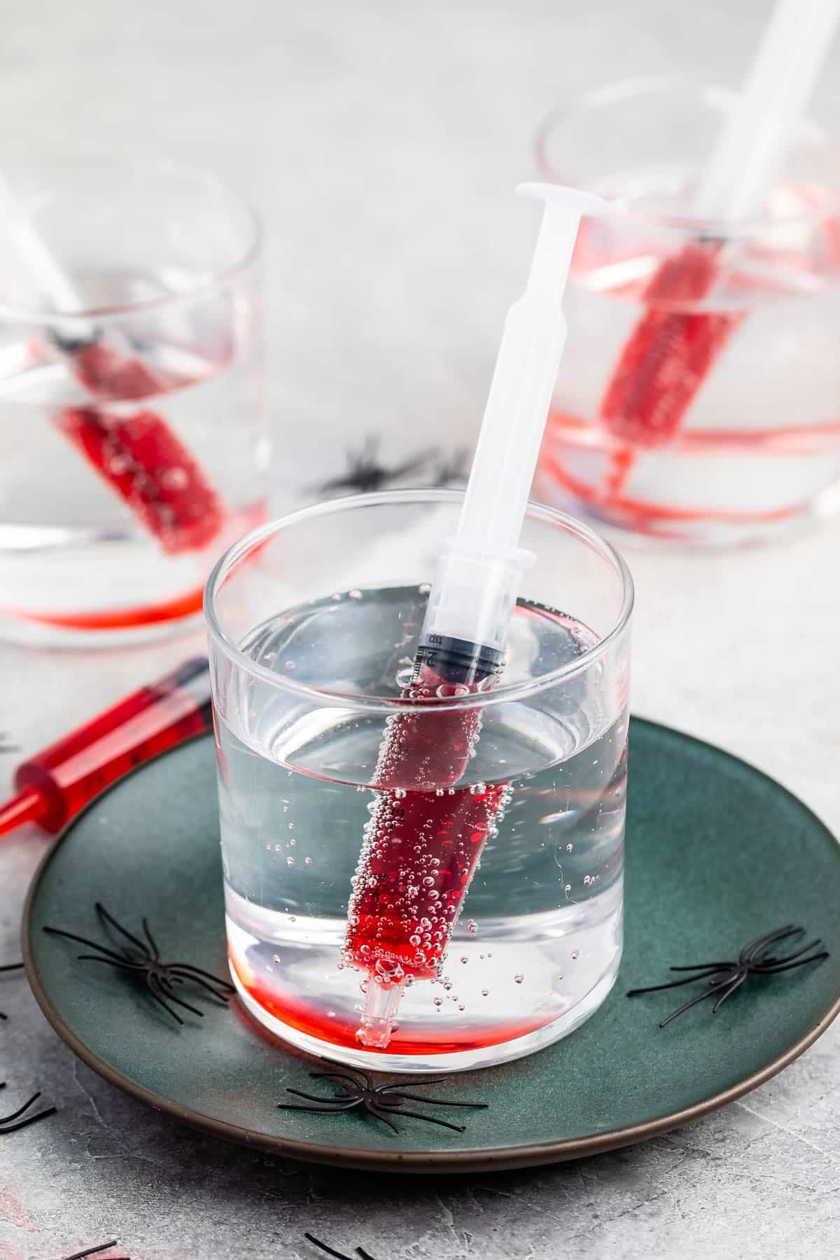 A clear drink with a syringe filled with red liquid inside the glass, served on a green plate with small plastic black spiders, and two more similar drinks in the background.