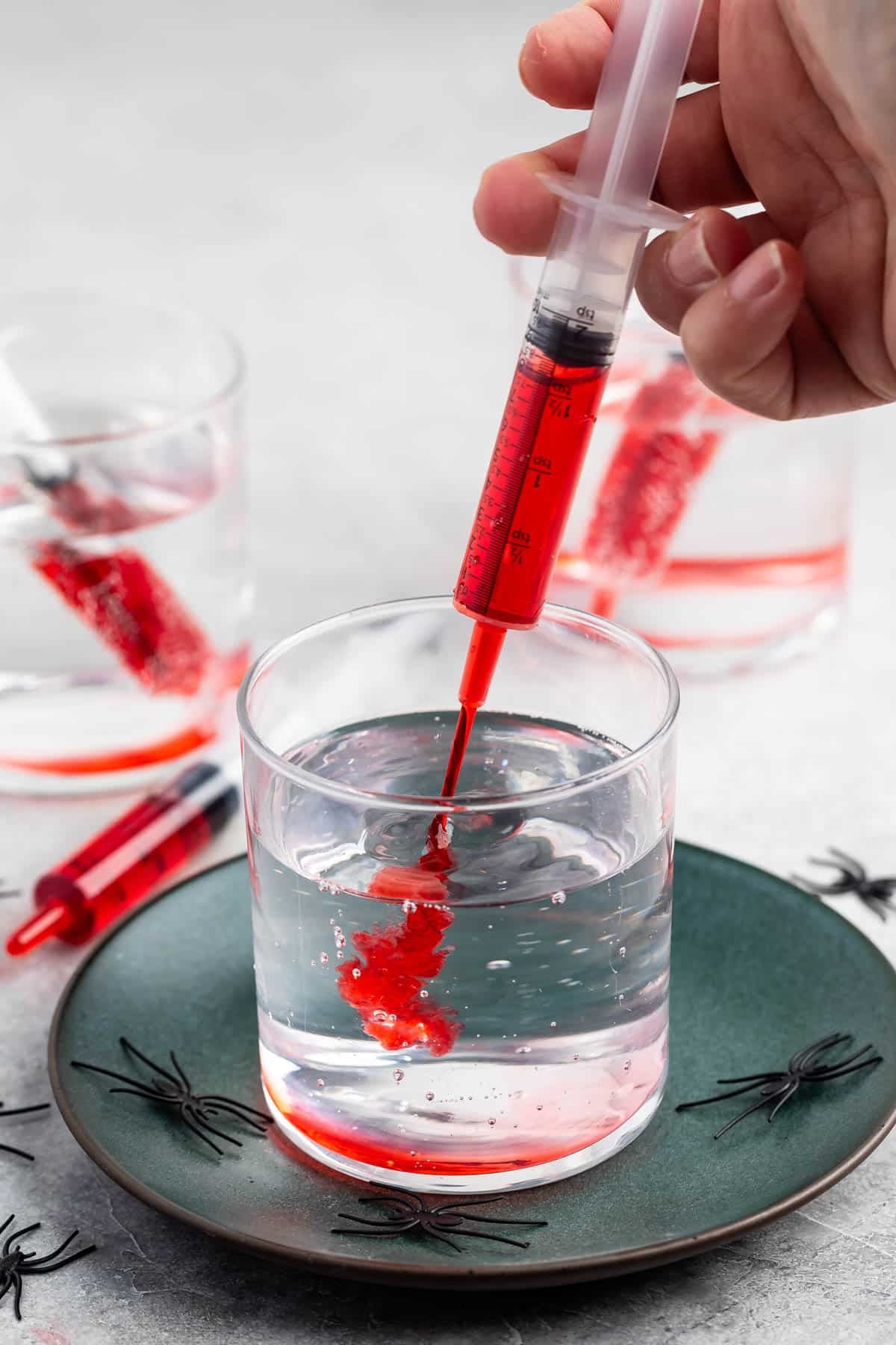 A hand injects red liquid from a syringe into a glass of clear water on a green plate, with plastic spiders scattered around and another glass with red liquid in the background.