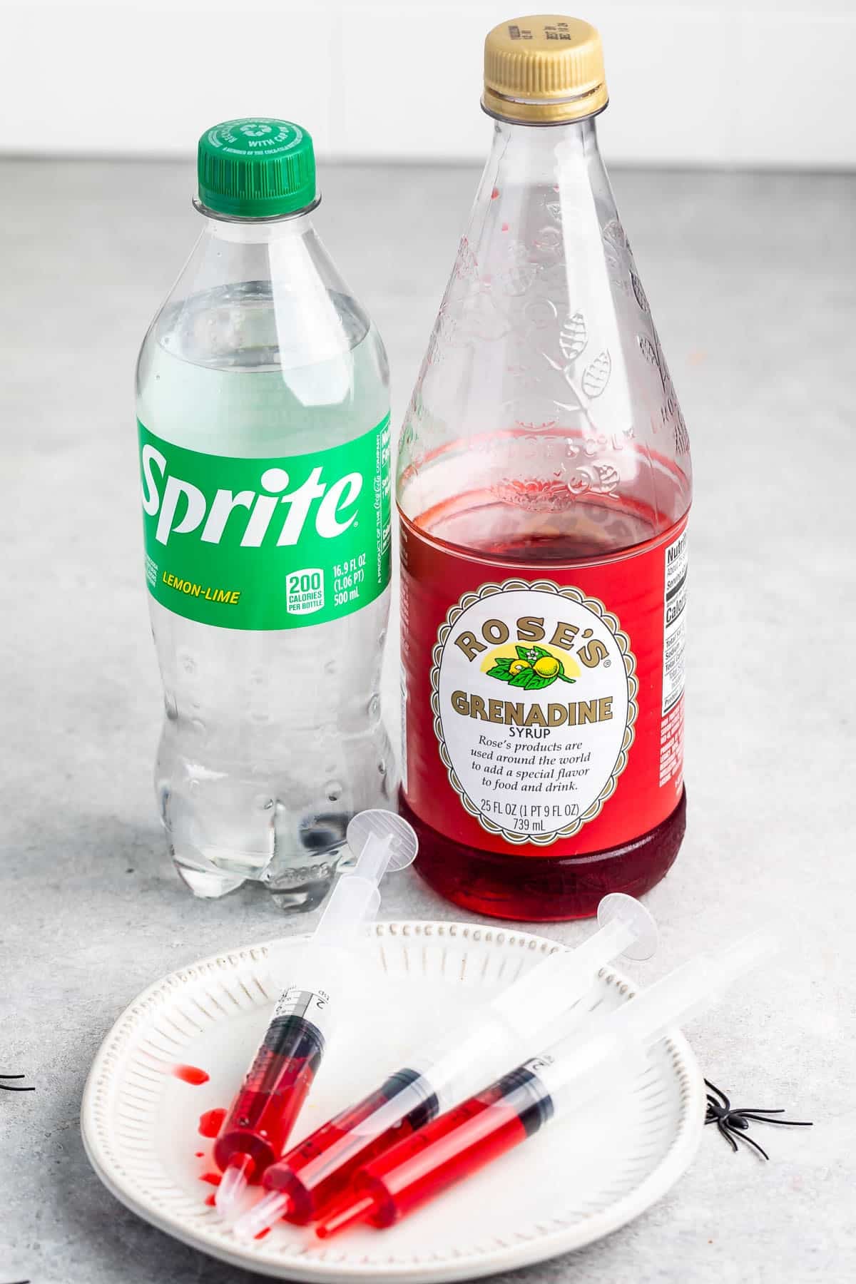 A bottle of Sprite, a bottle of Roses Grenadine syrup, and four plastic syringes filled with red liquid rest on a white plate.