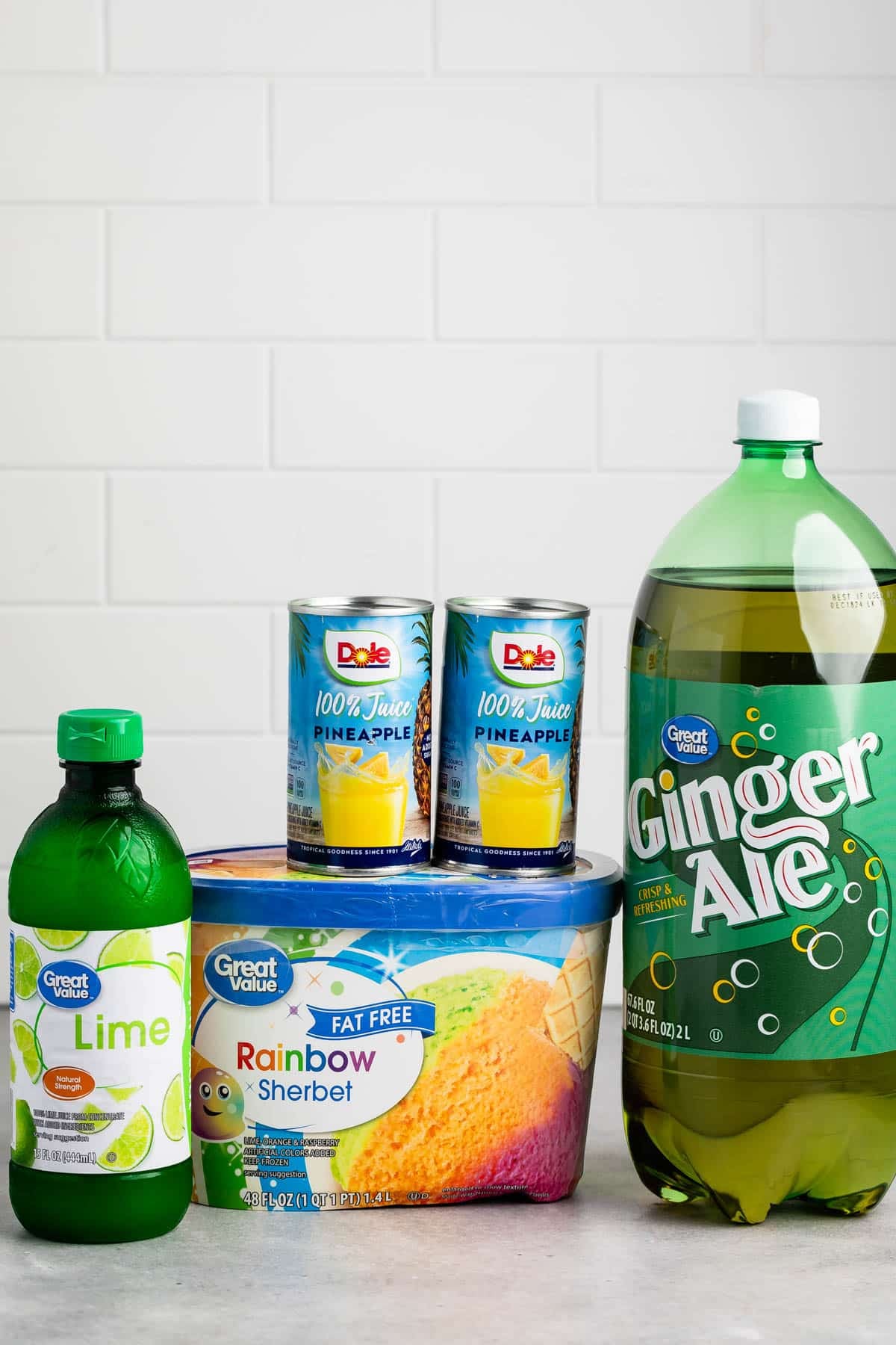 A bottle of lime juice, two cans of pineapple juice, a tub of rainbow sherbet, and a 2-liter bottle of ginger ale are arranged on a counter with a white tile background.