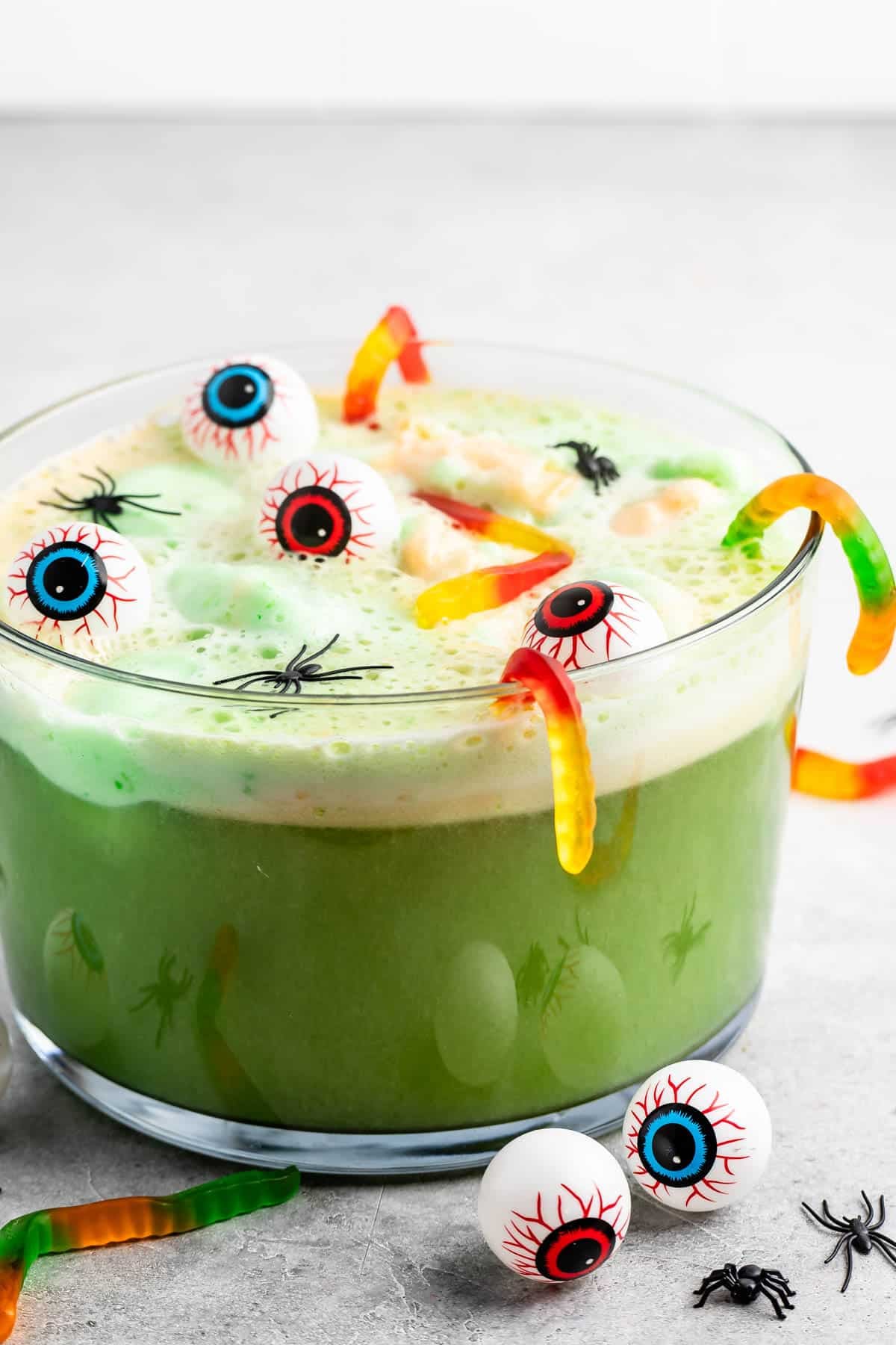 A glass bowl of green punch topped with floating fake eyeballs, gummy worms, and plastic spiders, creating a Halloween-themed drink.