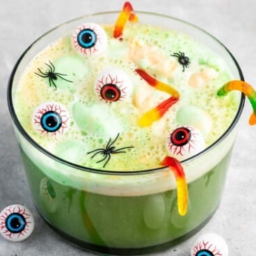 A green Halloween punch in a glass bowl with gummy worms, floating plastic eyeballs, and mini plastic spiders on top, surrounded by more eyeballs and spiders on the table.