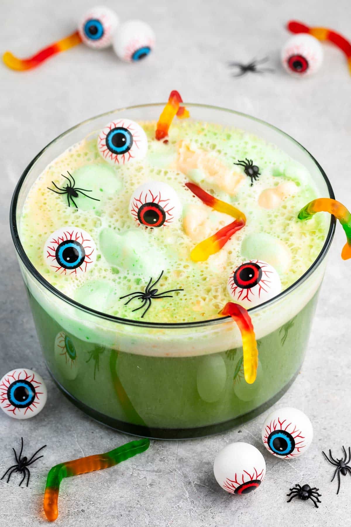 A green Halloween punch in a glass bowl with gummy worms, floating plastic eyeballs, and mini plastic spiders on top, surrounded by more eyeballs and spiders on the table.