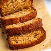 Three slices of moist banana bread are arranged on parchment paper in front of the remaining loaf. The bread has a golden-brown crust and a soft, crumbly texture.