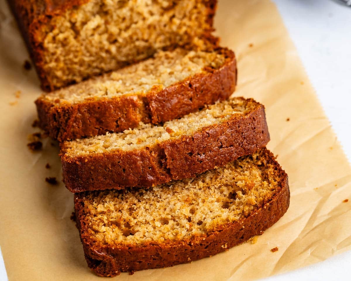 Three slices of moist banana bread are arranged on parchment paper in front of the remaining loaf. The bread has a golden-brown crust and a soft, crumbly texture.