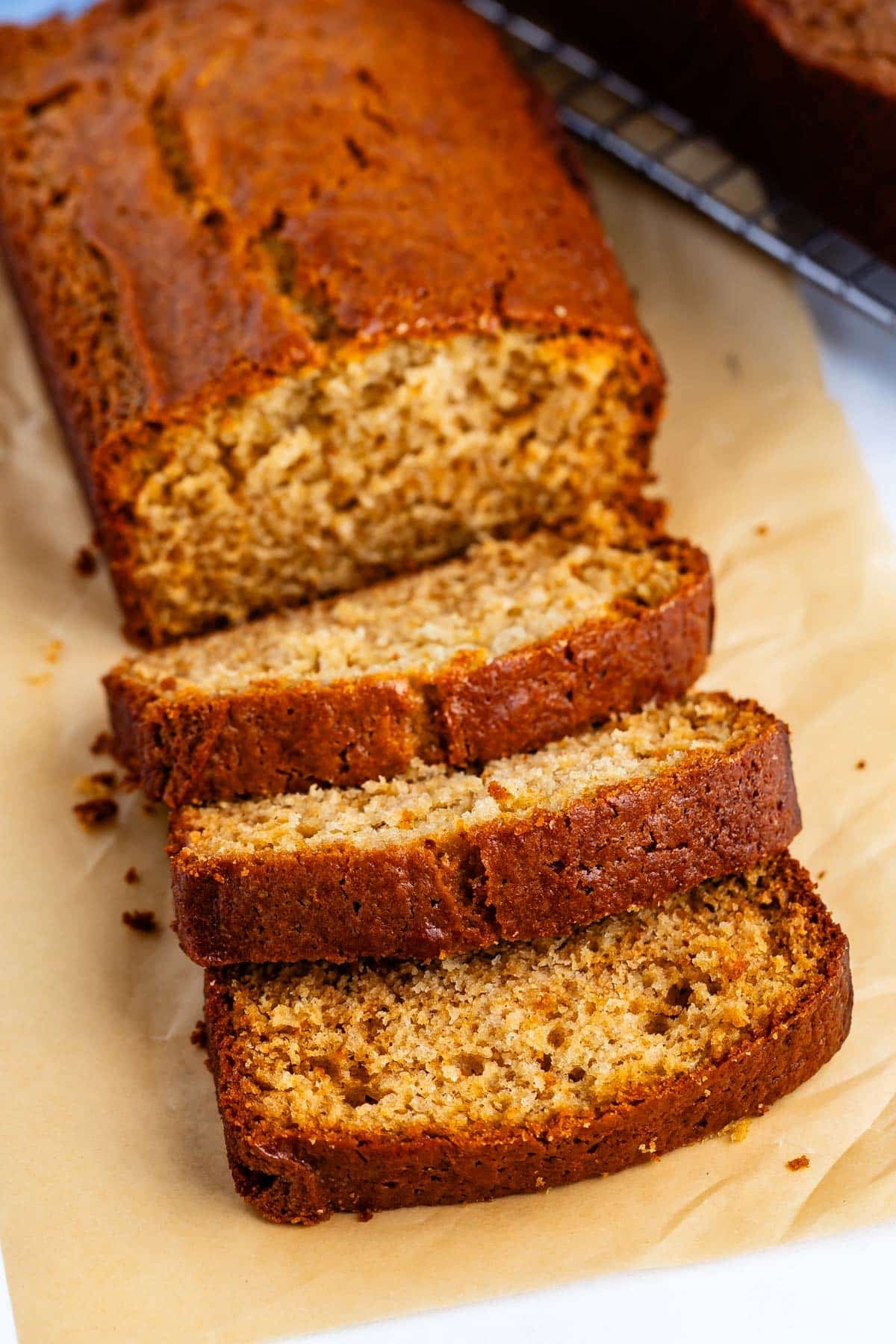 A loaf of banana bread on parchment paper, partly sliced with three pieces cut and fanned out in front of the loaf. The bread looks moist with a golden-brown crust.