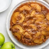 A caramelized upside-down apple cake sits on a white plate next to two green apples, with a napkin and a serving utensil nearby on a light-colored surface.