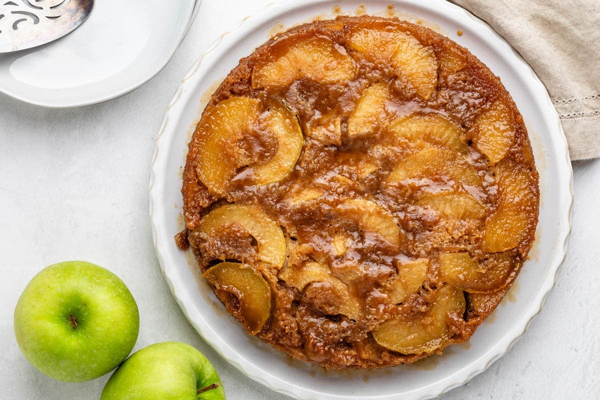 A caramelized upside-down apple cake sits on a white plate next to two green apples, with a napkin and a serving utensil nearby on a light-colored surface.