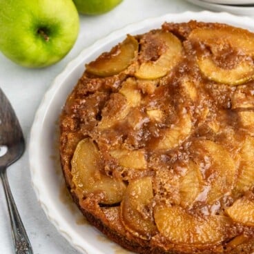 A caramelized apple upside-down cake on a white plate, with two green apples and a cake server nearby, set on a light surface.