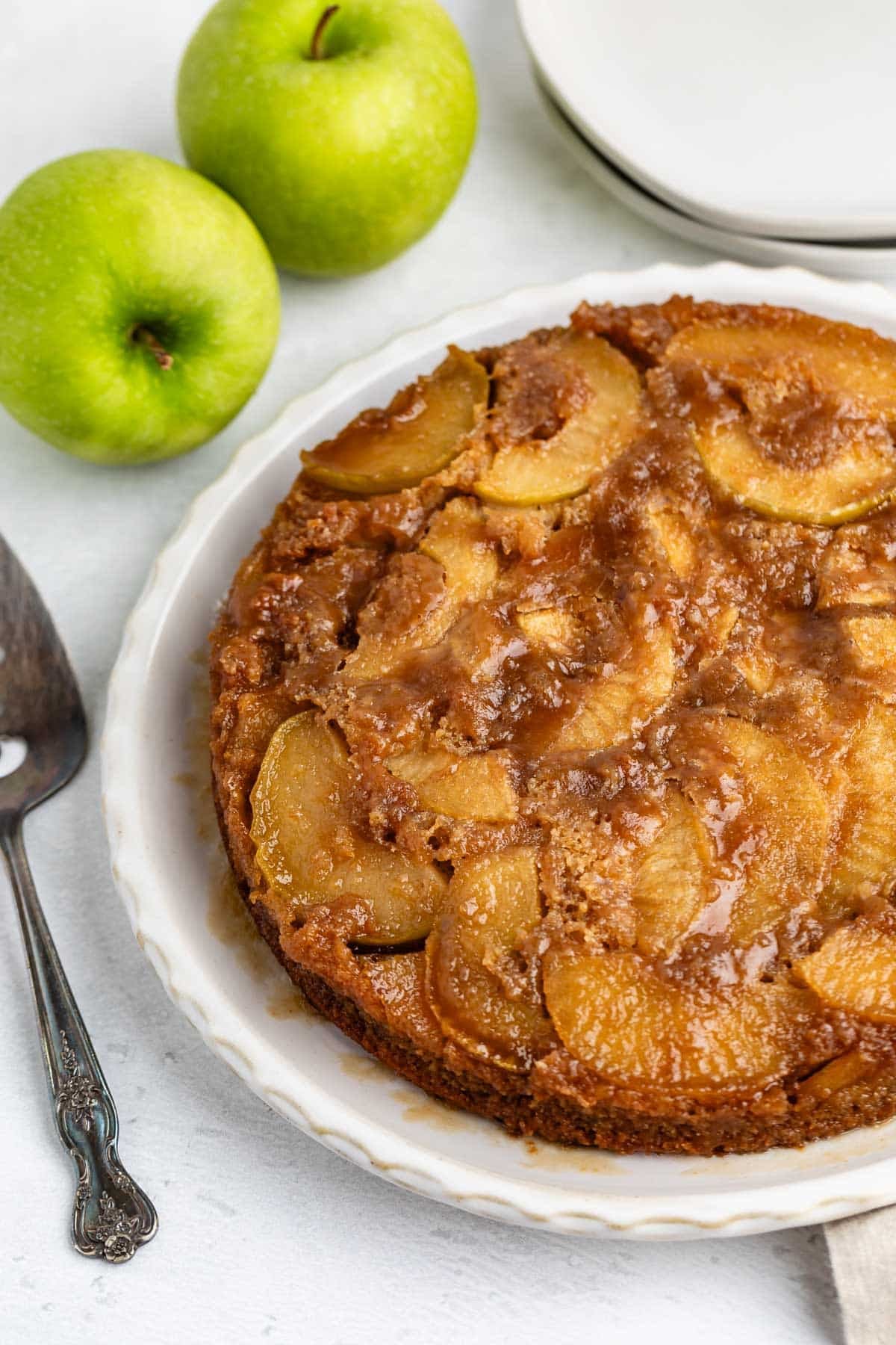 A caramelized apple upside-down cake on a white plate, with two green apples and a cake server nearby, set on a light surface.
